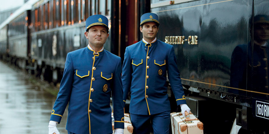 Two staff wearing uniform, walking down the station platform and carrying luggage with the Venice Simplon Orient Express behind them. 
